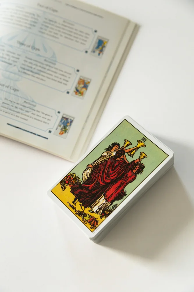 Carta Três de Copas do baralho Rider–Waite junto a um livro aberto.