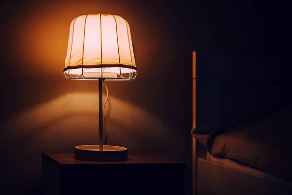 Abajur aceso sobre uma mesa de cabeceira, ao lado de uma cama, num quarto escuro.