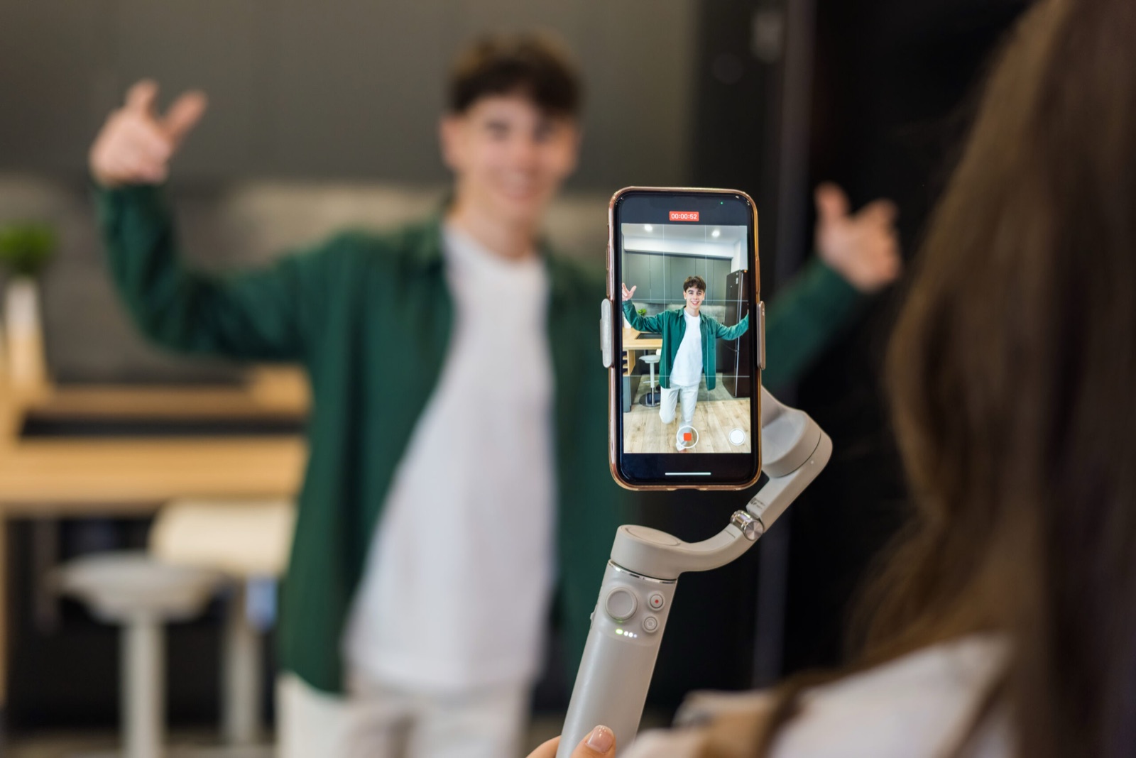 Jovem de camisa verde sorri de braços abertos para um celular preso a um estabilizador; na tela, o contador marca 52 segundos de gravação.